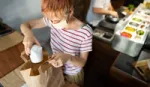 Woman kitchen worker packing food container into a takeaway bag with a chef preparing food in the background.