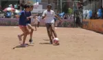 Girls playing football on an inner-city dirt pitch surrounded by fencing, being watched by a small group of people.