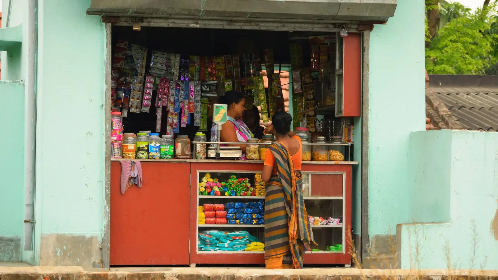 A small shop/kiosk with colourful products displayed on shelves, and a person wearing a traditional sari is standing at the counter.