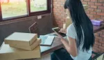 Asian woman at home online shopping. A laptop, an iPad and parcels surround her