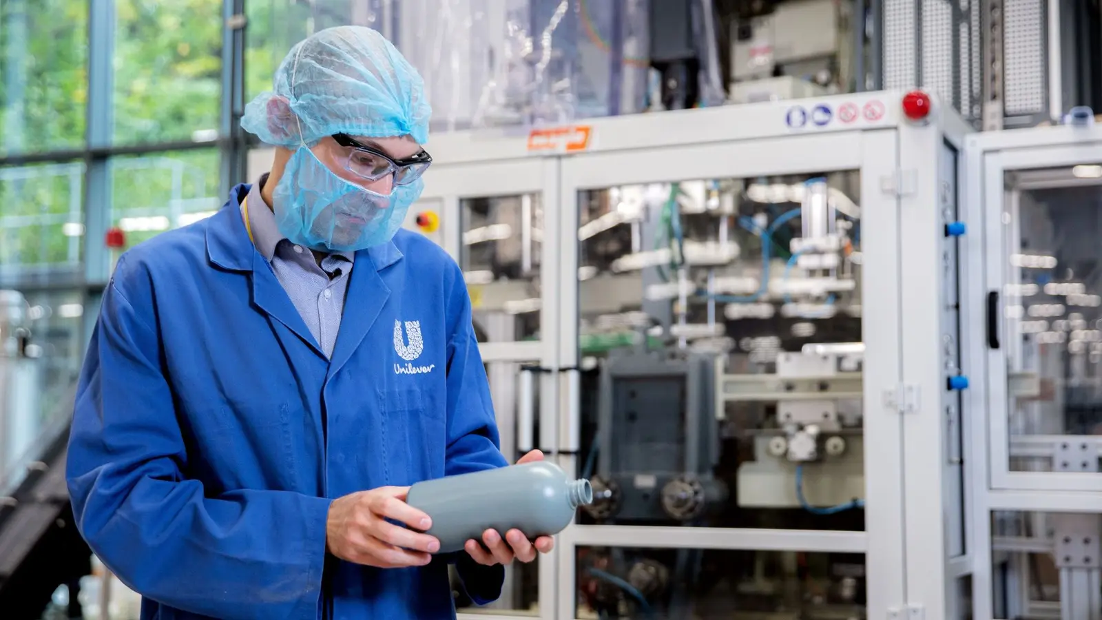 A person wearing a blue Unilever lab coat, hairnet, safety glasses and face mask examines a grey plastic bottle in a factory setting, with industrial equipment in the background.